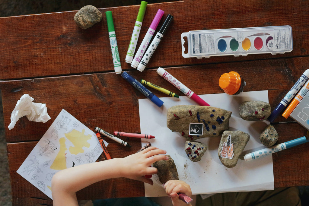 a child drawing at a table with markers, paints, and crayons